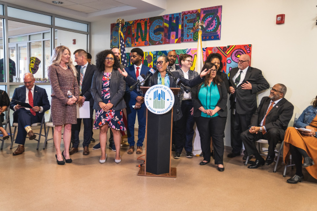 Eine Gruppe von Menschen steht vor einem Podium mit einem Mikrofon und einer Tafel mit der Aufschrift "Sancho City Councilman Joins City Council", während andere auf Stühlen sitzen oder in der Nähe stehen; Flaggen, ein Feuerlöscher und Deckenleuchten sind im Hintergrund zu sehen.