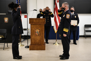 Gruppe von uniformierten Personen in Masken vor einem Podium mit Mikrofon, umgeben von Flaggen, Stühlen, Tischen und einer Wand, während einer Vereidungszeremonie, bei der eine Person ein Zertifikat hält.