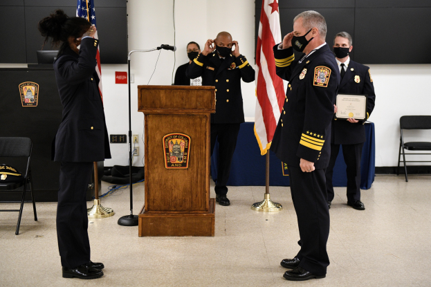 Gruppe von uniformierten Personen in Masken vor einem Podium mit Mikrofon, umgeben von Flaggen, Stühlen, Tischen und einer Wand, während einer Vereidungszeremonie, bei der eine Person ein Zertifikat hält.