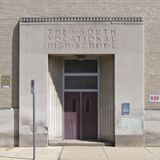 Das Südberufsschulgebäude mit einer Tür, Schildern an der Wand und einem Weg, der zur Eingangstür führt.