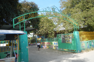 Der Eingang einer Schule mit einem grünen Torbogen, einem Metallzaun, einer Fahne mit Bildern und Text, einer Gruppe von Bäumen, nahegelegenen Häusern mit Dächern und einer Person, die auf dem Boden unter einem bewölkten Himmel steht.