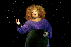 Eine Frau in einem lilafarbenen Kleid steht auf einer Bühne, beleuchtet von ein paar Lichtern im Hintergrund, als ob sie mitten in einer Vorstellung ist.