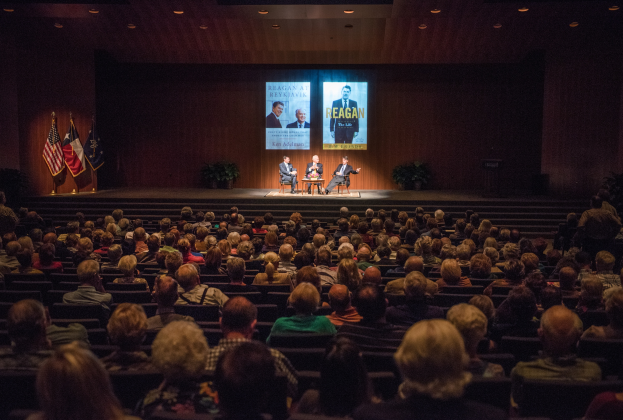 Eine große Menschenmenge sitzt vor einer Bühne mit drei Personen in Stühlen im Vordergrund, drei Fahnen auf der linken Seite und einem Bildschirm mit Text und Bildern im Hintergrund, unter einer Decke mit Beleuchtung.