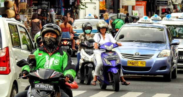 Eine Gruppe von Menschen, die Motorräder auf einer belebten Straße fahren, mit Autos und anderen Fahrzeugen, einige tragen Helme, vor Gebäuden, Bäumen, Schildern und Geschäften.