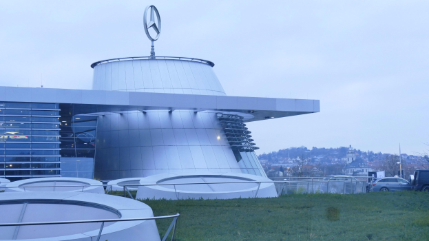 Das Mercedes-Benz Museum in Frankfurt, Deutschland, mit seinem Logo auf einem große Geböude mit Gras, Gelöndern, Fahrzeugen auf der Straße, Böumen im Hintergrund und einem klaren blauen Himmel.