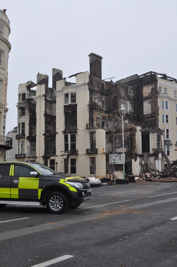 Ein vor einem schwerbeschädigten Gebäude geparkter Polizeiwagen mit zersplitterten Fenstern und verstreuter Trömmern, unter einem sichtbaren Himmel mit umliegenden Gebäuden.