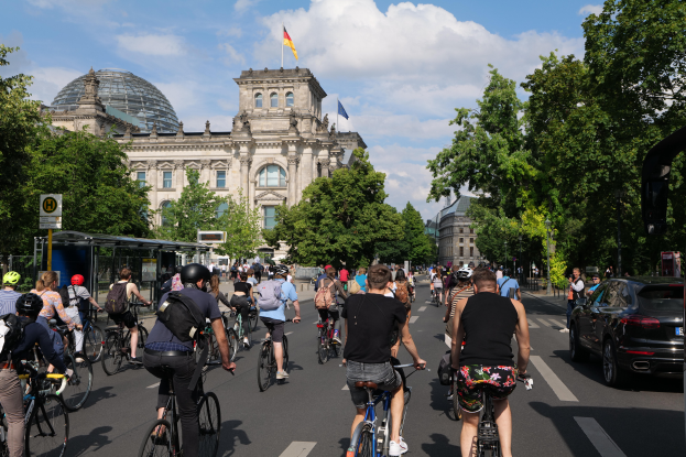 Eine Gruppe von Menschen fährt mit Fahrrädern eine von Bäumen gesäumte Straße in Berlin, Deutschland, entlang, mit Gebäuden auf beiden Seiten, einem Bushaltestellen auf der rechten Seite und einer Flagge, die auf einem der Gebäude weht, unter einem bewölkten Himmel.