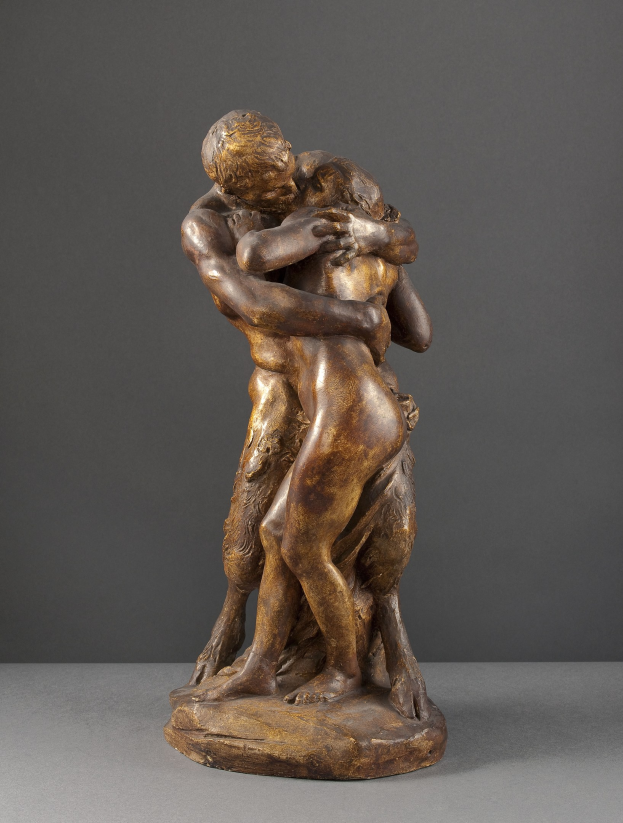 Bronzestatue mit dem Titel "Der Kuss" von Jean-Baptiste-Camille Corot, die einen Mann in einem weißen Hemd und blauen Jeans zeigt, der eine Frau in einem orangen Kleid umarmt, mit detaillierten, realistischen Details und einem warmen goldenen Ton gegen einen grauen Hintergrund.