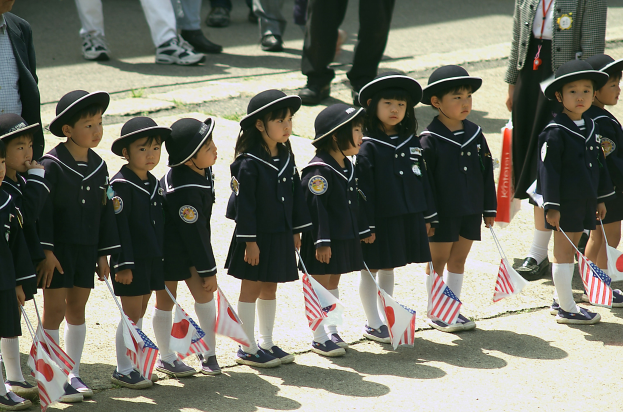 Eine Gruppe von Kindern in Schuluniformen, die nebeneinander stehen und jeweils eine Fahne halten, mit ein paar Menschen im Hintergrund auf einer Straße.
