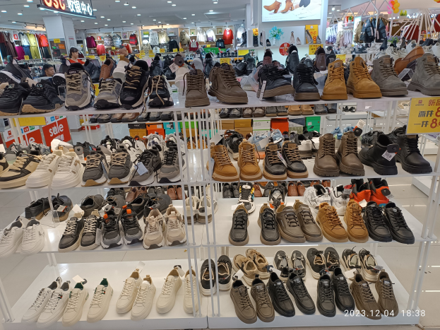 Ein Schuhgeschäfts-Innenraum mit verschiedenen Schuhen auf Gestellen, Preisetiketten sichtbar, Menschen im Hintergrund, hängende Kleidung, Deckenlampen, ein Bildschirm und Texttafeln.