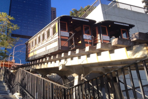 Gebäude im Hintergrund, ein Baum links, eine Treppe mit Geländer unten und eine Bahnlinie mit zwei Zügen rechts.