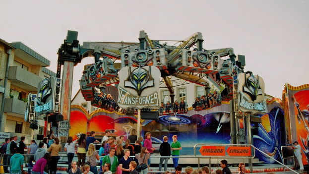Menschenmenge um eine Karussellfahrt mit einigen darauf sitzend und anderen auf Treppen, umgeben von Geländern und Texttafeln, mit Gebäuden und einem klaren blauen Himmel im Hintergrund.