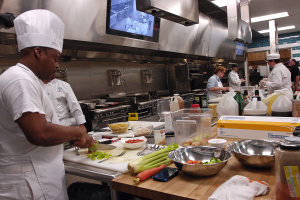 Eine Gruppe von Köchen in einer Küche, die Essen auf einem Schneidbrett zubereiten, wobei ein Koch in der foreground in einer weißen Schürze und einer weißen Mütze steht und ein Messer hält und von verschiedenen Utensilien, Schüsseln, Gemüse, einem Mobiltelefon, Flaschen und anderen Gegenständen umgeben ist, mit einem Fernseher und Deckenlampen im Hintergrund.