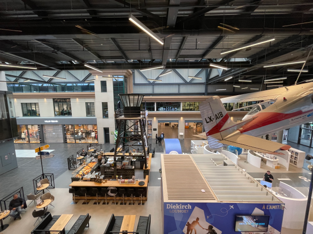 Ein geräumiger Raum mit zahlreichen Tischen, Stühlen, Sofas und verschiedenen Gegenständen, darunter einige Personen, mit einem Flugzeug auf der rechten Seite und Glasfenstern und -wänden im Hintergrund.