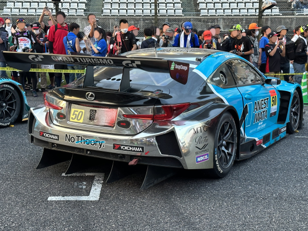 Ein schwarzer Lexus RC F GT3 Rennwagen umgeben von Menschen in Mötzen und Schutzbrillen, einige halten Kameras und Handys, mit Maschendrahtzaun und verstreuten Stühlen im Hintergrund.