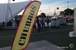 Ein buntes Faschingszelt mit Zelten, einem zentralen Mast, Menschen, Bäumen, Strommasten, Straßenbahnen auf der Straße und einem Eisenständer links.