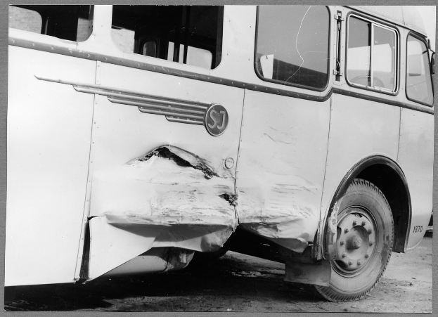 Schwarzes und weißes Foto eines beschädigten Busses am Straßenrand mit eingedellter Front, zersplitterten Fenstern und geöffneter Fahrertür, die den Innenraum zeigt.