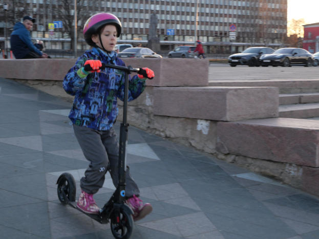 Ein junger Junge in Helm und Handschuhen fährt mit einem Scooter eine Treppe hinunter, vorbei an Autos, Menschen, Bäumen, Pfosten, Brettern, Gebäuden und einem klaren blauen Himmel.