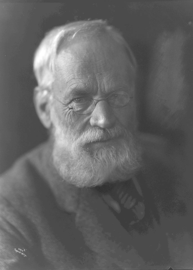 Alter Mann mit Vollbart und Brille, trägt dunklen Anzug, schaut ernst in die Ferne