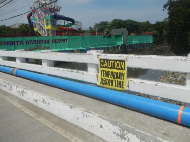 Vorsichtsschild mit der Aufschrift "Vorsicht temporäre Wasserleitung" auf einer Brücke mit Geländer, Bäumen, Gebäuden, einer Wasserrutsche und einem bewölkten Himmel im Hintergrund.