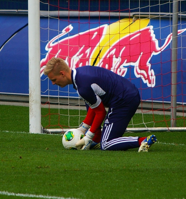 Ein Fussballspieler kniet auf dem Boden mit einem Ball vor einem Tor, umgeben von Gras und einem Netz, mit einem Banner im Hintergrund mit Text und einem Bild einer Person.