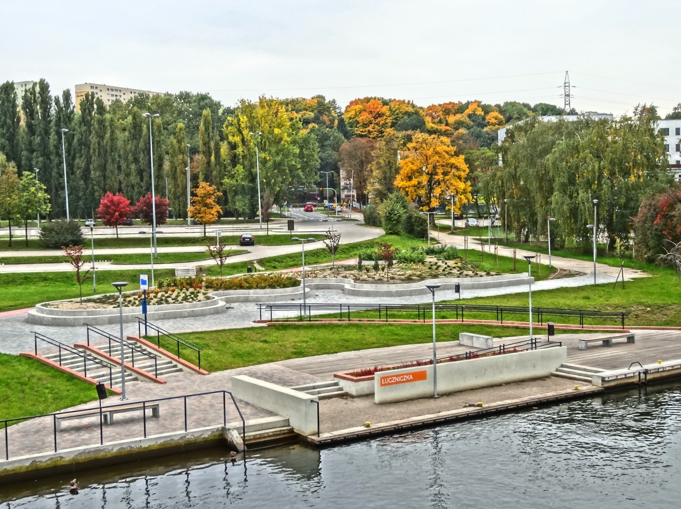 Ein Fluss fließt durch einen grünen Park neben hohen Gebäuden mit Bäumen, Straßeninfrastruktur, Fahrzeugen und einem bewölkten Himmel.