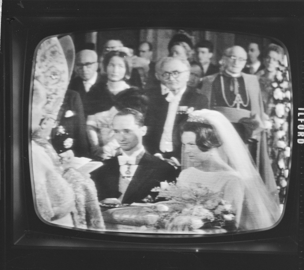Schwarzes und weißes Foto einer Braut in einem weißen Kleid und Schleier, die einen Strauß hält, neben einem Bräutigam in einem Anzug und Krawatte, auf einem Fernsehbildschirm umgeben von einer Gruppe von Menschen angezeigt.