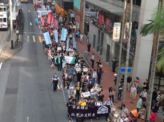 Ein großer Protestmarsch in Hong Kong mit Menschen, die Schilder und Plakate tragen, flankiert von Bäumen und Gebäuden auf der rechten und Fahrzeugen auf der linken Seite.