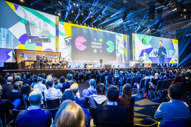 Eine große Menschenmenge sitzt auf Stühlen vor einer Bühne bei der Deutschen Internetkonferenz 2019, mit einigen Personen auf dem Podium im Vordergrund, einem Tisch mit verschiedenen Gegenständen links und Bildschirmen und Lichtern im Hintergrund.