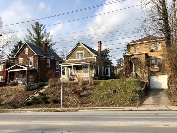 Reihe von Häusern mit Fenstern, Türen, Säulen, Geländern, Stufen, Pflanzen und einem Pfahl mit einer Tafel entlang einer Straße, vor einem bewölkten Himmel.