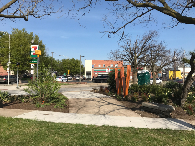 Eine städtische Straße mit parkenden Autos, Grünflächen, Straßenausstattung und Gebäuden, mit einem 7-Eleven-Parkplatz im Vordergrund und Himmel im Hintergrund.