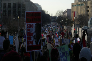 Menschen mit Schildern auf der Straße protestierend, mit Gebäuden, Bäumen, Laternenmästen und Himmel im Hintergrund.