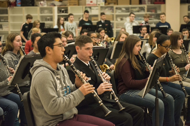 Eine Gruppe von Menschen, die Klarinetten in einem Klassenzimmer spielen, mit Notenpulten, einige sitzen und einige stehen, mit Regalen voller Kisten im Hintergrund.