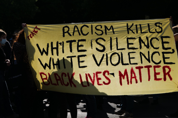 Eine Gruppe maskierter Personen steht nachts auf einer Straße und hält ein Schild mit der Aufschrift "Rassismus tötet Weiße Stille, weiße Gewalt, Schwarze Leben zählen".