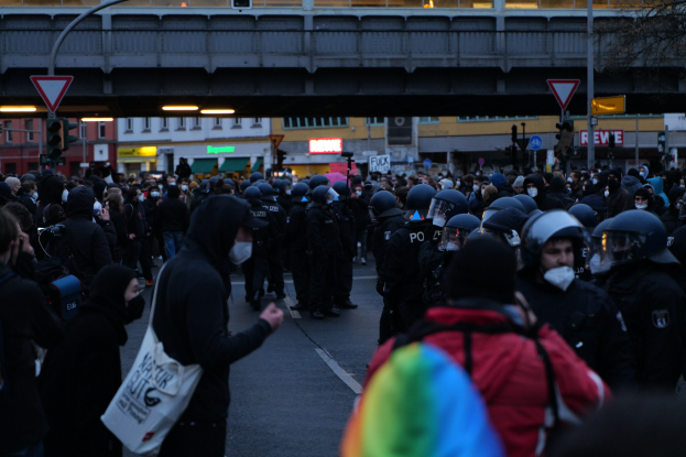 Eine große Gruppe von Menschen steht vor einer Menge von Polizisten, einige tragen Helme und tragen Taschen oder halten Kameras, mit Gebäuden, Schildern, Laternenmasten, Verkehrsampeln, einer Brücke und einem Baum im Hintergrund.