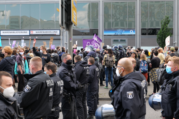 Eine große Gruppe von Menschen steht vor einem Gebäude, einige halten Schilder und tragen Helme, mit einem Schilderpfahl im Vordergrund und einem Baum im Hintergrund, was auf eine Protestaktion hindeutet.