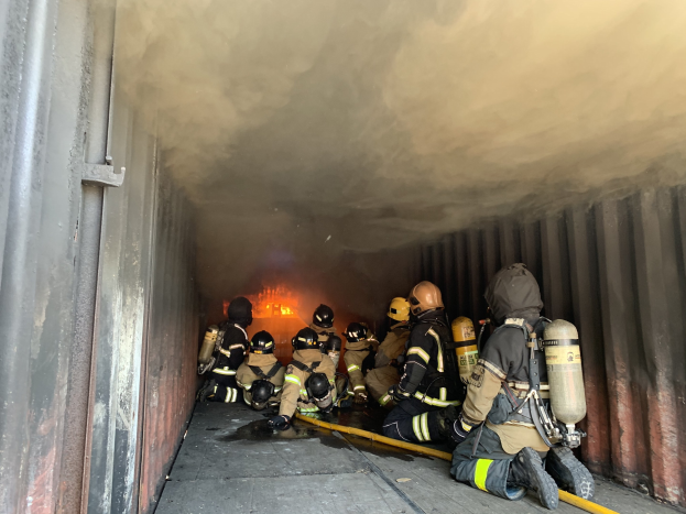 Feuerwehrleute in Schutzausrüstung arbeiten in einem raucherfüllten Tunnel, einer hält einen Schlauch, mit Flammen im Hintergrund.