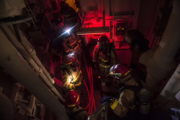 Feuerwehrleute in Helmen und Sauerstoffflaschen stehen in einem dämmrig beleuchteten Raum mit Rohren an den Seiten und bereiten sich auf einen Rettungseinsatz in der Nähe von Maschinen im Hintergrund vor.