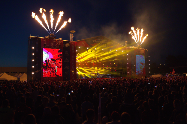 Eine große Menschenmenge versammelt sich auf einem Musikfestival, mit einer Bühne in der Mitte, auf der Musiker von hellen Scheinwerfern beleuchtet werden und von Bannern umgeben sind, Zelte links und ein dunkler Himmel mit Feuerwerk im Hintergrund.