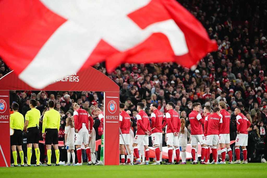 Eine Gruppe von Menschen auf einem Fußballfeld mit einer roten und weißen Flagge im Vordergrund, einem Bogen mit der Aufschrift "Bayern München vs Bayern München Wetten & Vorschau" im Hintergrund und einer großen Stadionmenge.