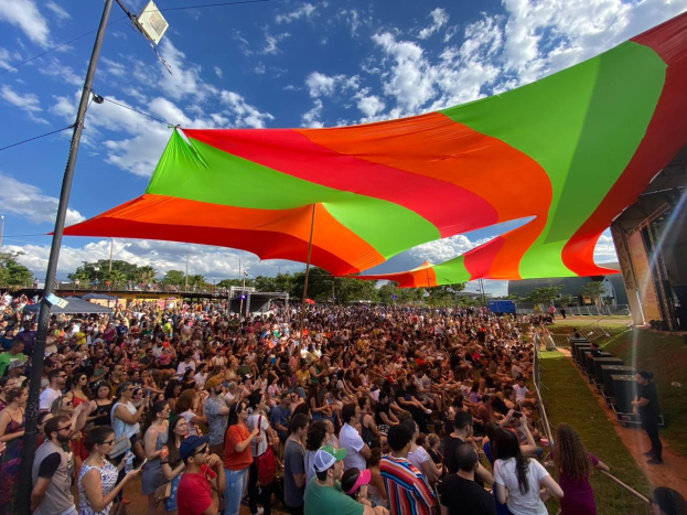 Eine große Menschenmenge steht vor einer Regenbogenflagge, mit einem Gebäude, einer Bühne, Lautsprechern, Bäumen und Wolken im Hintergrund, die sich zu einem Festivaljubiläum versammelt hat.