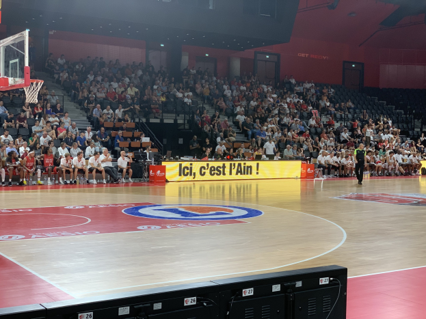 Ein Basketballfeld in einer Turnhalle mit Zuschauern auf den Rängen, ein Basketballkorb links und elektronische Geräte unten im Bild.