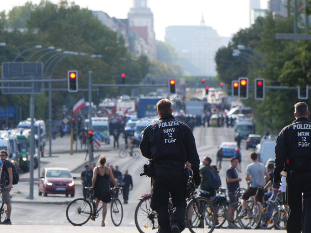 Zwei Polizeibeamte stehen an der Seite einer Straße neben einer Gruppe von Menschen auf Fahrrädern, mit Fahrzeugen, Verkehrszeichen, Bäumen, Gebäuden und einem klaren blauen Himmel im Hintergrund.