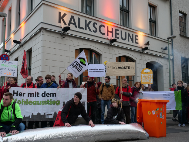 Eine Gruppe von Menschen protestiert vor einem Gebäude in Deutschland, hält Schilder und Plakate hoch, mit zwei Personen im Vordergrund und einem Müllcontainer auf der rechten Seite.