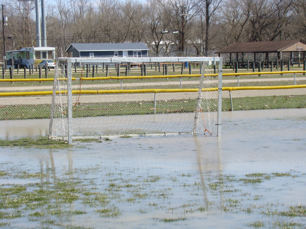 Ein Fußballtor steht in der Mitte eines überfluteten Feldes mit umgebenem Gras und Wasser, mit Hütten, Pfählen, Bäumen, Fahrzeugen und einem klaren blauen Himmel im Hintergrund.