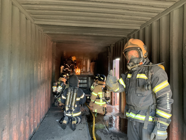 Feuerwehrleute in Schutzausrüstung innerhalb eines Containers, einer hält ein Rohr, mit einem Feuerlöscher im Hintergrund.