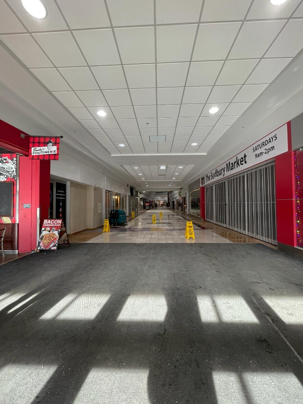 Innenansicht eines Einkaufszentrums mit Deckenbeleuchtung, einem Stuhl und einer Tafel mit der Aufschrift "Sydbury Market - Vorher und Nachher" auf der linken Seite und verstreuten Gegenständen auf der rechten Seite und auf dem Boden.