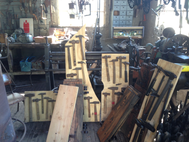 Ein mit verschiedenen Werkzeugen, Geräten und Holzplanken gefüllter Werkstattraum, mit einem Stuhl links, Werkzeugen an der Wand und einem Fenster im Hintergrund.