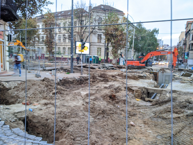 Baustelle mit einem großen Loch, das von einem Metallzaun umgeben ist, umgeben von Fahrzeugen, Fußgängern, Verkehrsampeln, Schildern, Bäumen, Gebäuden und einem klaren blauen Himmel.