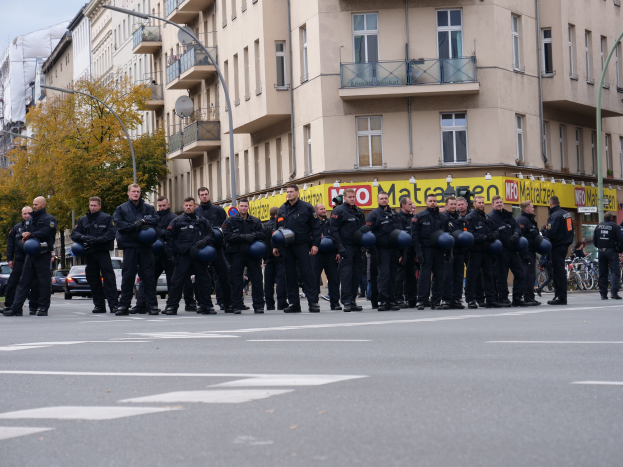 Eine Gruppe von Polizisten in schwarzen Uniformen mit blauen Helmen steht in einer Straße, die von Laternenpfählen, Bäumen und Glasfenstergebäuden gesäumt ist, unter einem klaren blauen Himmel.
